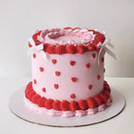 A round cake with pink and red decorative icing, featuring heart shapes and white ribbon details.