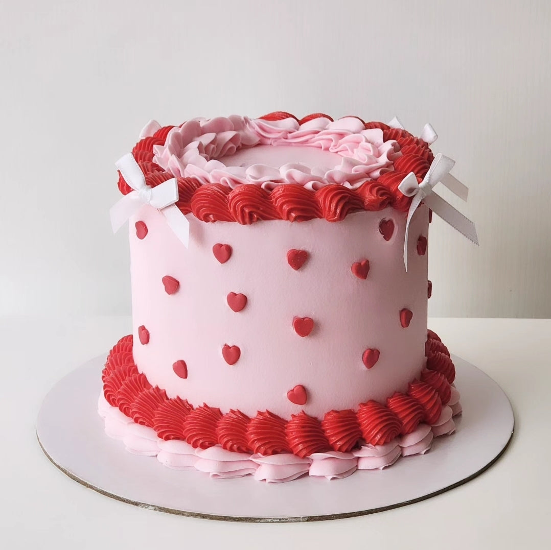 A round cake with pink and red decorative icing, featuring heart shapes and white ribbon details.
