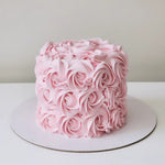 A multi-layered pink cake decorated with swirls of creamy white frosting and vanilla buttercream, placed on a white cake stand against a neutral background.