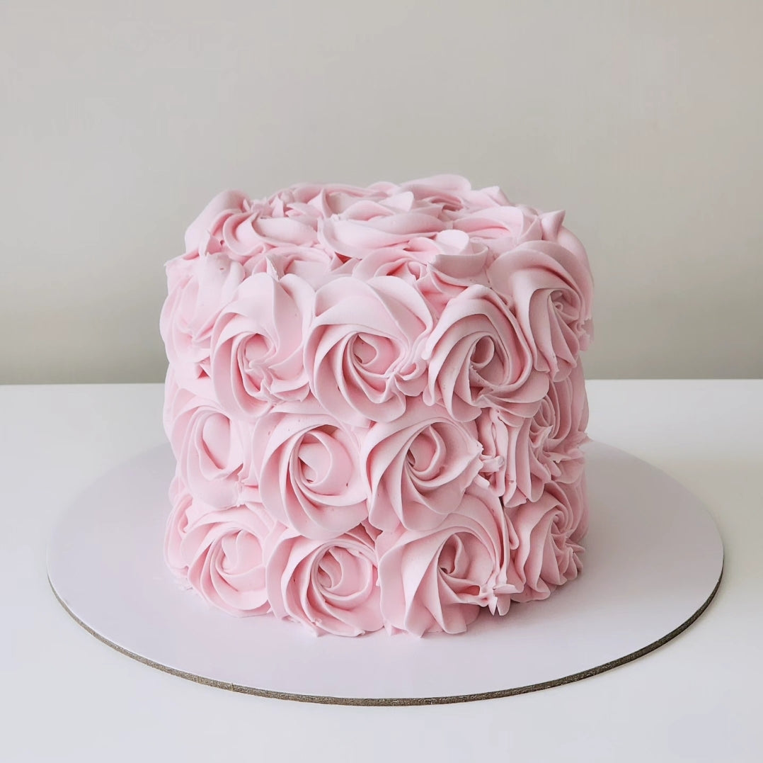 A multi-layered pink cake decorated with swirls of creamy white frosting and vanilla buttercream, placed on a white cake stand against a neutral background.