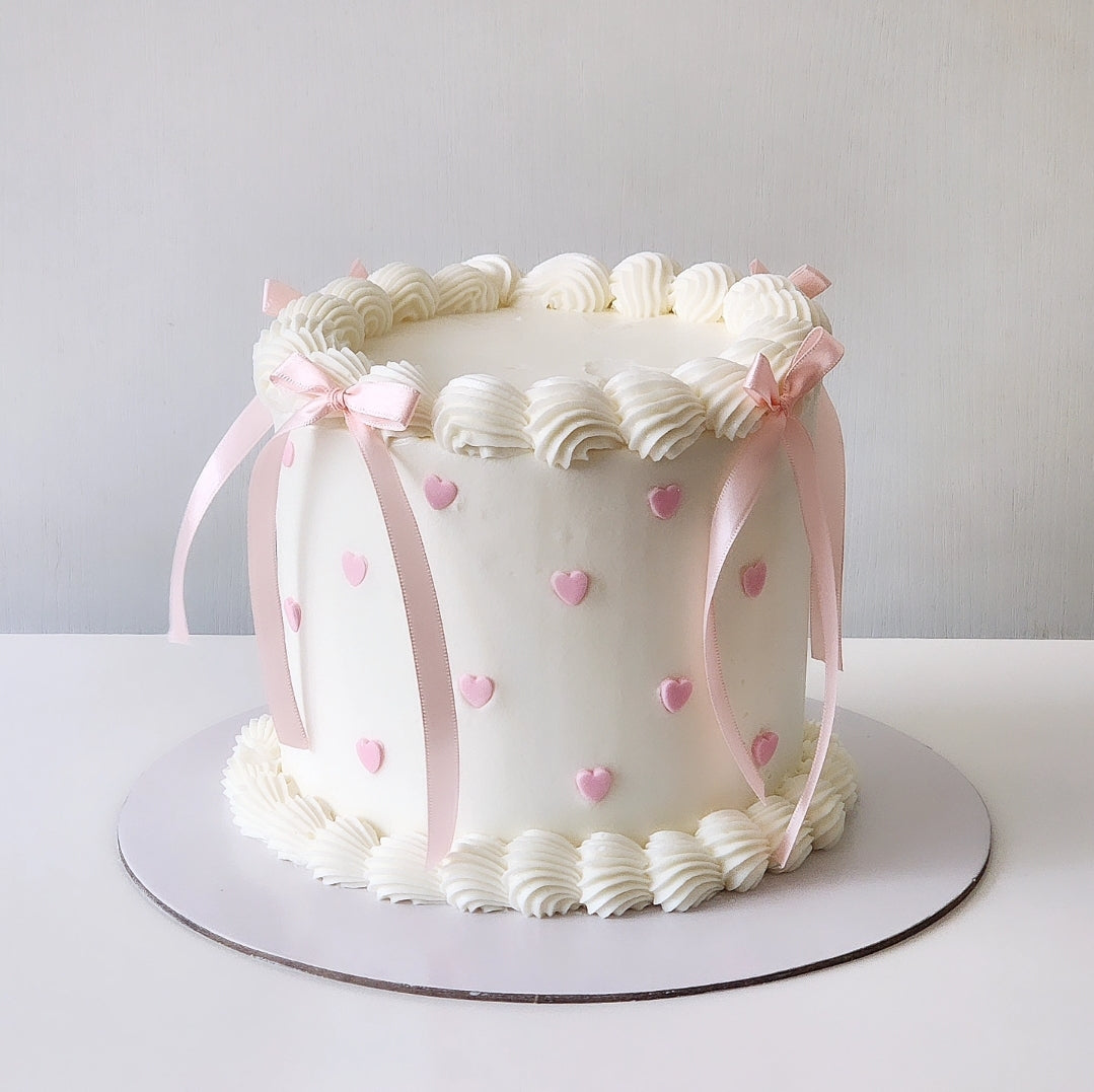 A decorated cake with white frosting and pink accents, featuring a layer of white piped swirls and a satin black ribbon around the base.
